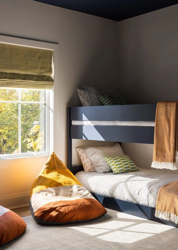 A bright, cozy children's bedroom with a dark navy bunk bed, patterned bedding, mustard and rust beanbag seating, and warm tan throws. Sunlight streams through a window with a woven Roman blind, creating strong geometric shadows across the carpet and bed.