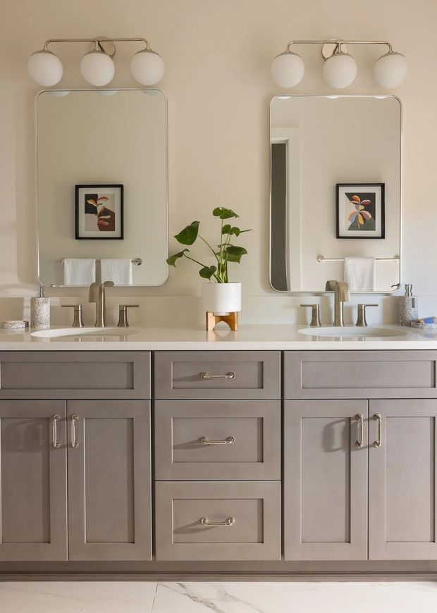 A symmetrical, double-sink bathroom vanity with taupe-grey shaker-style cabinetry, a pale stone countertop, brushed metal fixtures, twin rounded-rectangle mirrors, and three-globe wall lights above each mirror. Framed botanical artwork, white towels, and a potted plant add understated decorative detail. The room has a refined, warm contemporary feel.