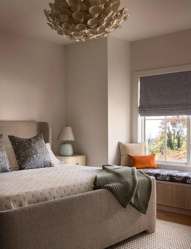 A softly lit, refined bedroom featuring a fully upholstered beige bed, patterned decorative pillows, a textured bedspread, and a muted green throw. A sculptural clustered pendant light hangs from the ceiling, while a small bedside table with a pale lamp sits beside the bed. The room includes a window seat with cushions, a grey Roman blind, and warm neutral wall and flooring finishes.