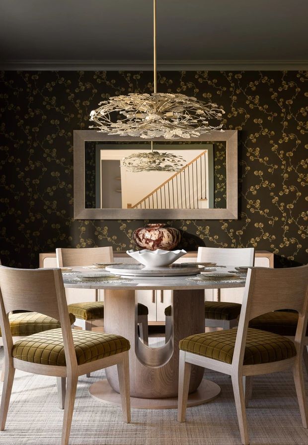 A refined dining room with a round sculptural timber dining table surrounded by upholstered timber dining chairs. The room features dark floral wallpaper, a large rectangular wall mirror, a dramatic suspended botanical chandelier, and layered neutral furnishings with olive-green seating accents.