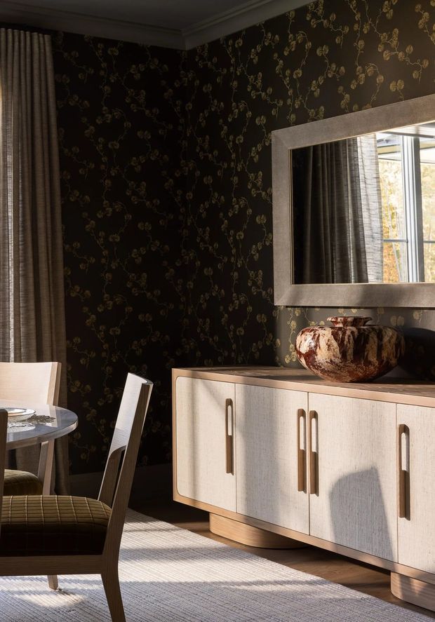 A richly styled dining area with dark floral-patterned wallpaper, tailored drapery, a round dining table and upholstered chairs, and a light wood sideboard with elongated handles. A large framed mirror reflects the window and curtains, while warm sunlight creates strong shadows across the textured rug and cabinetry. The overall look is refined, moody, and mid-century influenced.