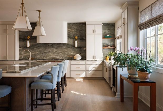 A refined, spacious kitchen with a large island and integrated seating, white raised-panel cabinetry, a dramatic dark veined stone backsplash, and warm timber flooring. Soft natural light enters through tall windows with woven Roman shades, while sculptural pendant lights and potted orchids add decorative detail.
