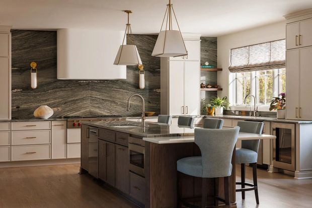 A spacious, refined kitchen with a large dark wood island, light cabinetry, a dramatic veined stone backsplash, integrated appliances, and a peninsula with upholstered bar stools. Two sculptural pendant lights hang over the island, while a bright windowed breakfast/work area and open shelving add warmth and functionality.