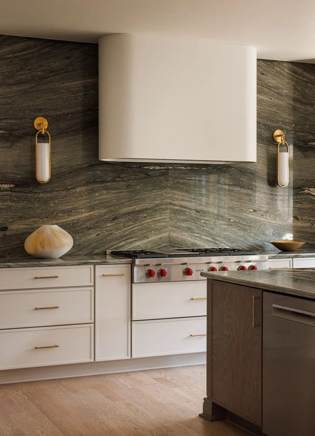 A refined kitchen featuring a large professional stainless-steel range beneath a sculptural white range hood. The walls are clad in dark, richly veined stone forming a dramatic backsplash, contrasted with pale cabinetry, brass hardware, warm wood flooring, and a dark wood island with a stone benchtop. Decorative wall sconces and understated accessories add a luxurious, tailored feel.