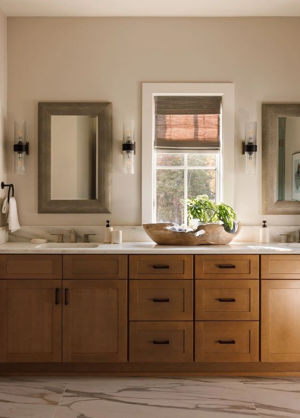 A warm, traditionally styled bathroom vanity with extensive medium-toned wood cabinetry, a pale stone countertop with integrated sinks, framed mirrors, wall sconces, and a central window with layered woven blinds. A sculptural wooden bowl with greenery sits on the counter, and large-format veined stone tiles cover the floor.