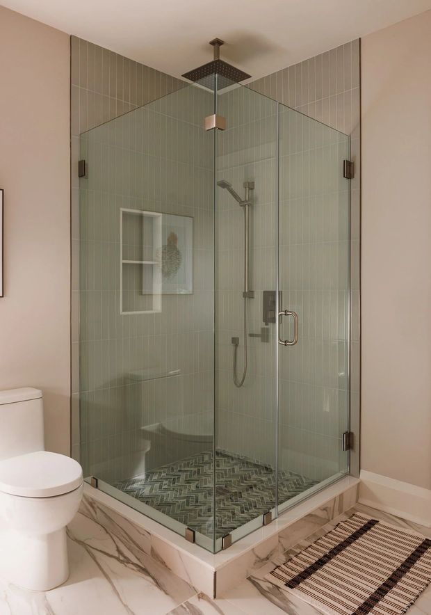 A contemporary bathroom featuring a large corner glass-enclosed shower with pale green vertically stacked wall tiles, a square rainfall showerhead, handheld shower, and dark metal fixtures. The shower base and bathroom floor use marble-look tiles, with a built-in shower niche and a striped floor mat. A white toilet is visible in the foreground.