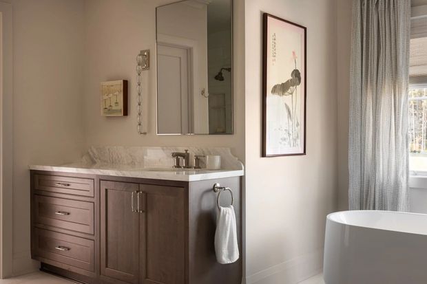 A warm, refined bathroom featuring a dark wood vanity with drawers and cabinet doors, a light veined stone benchtop and integrated basin, brushed metal tapware, a large mirrored cabinet, framed botanical artwork, and a freestanding white bathtub beside a window with sheer curtains.
