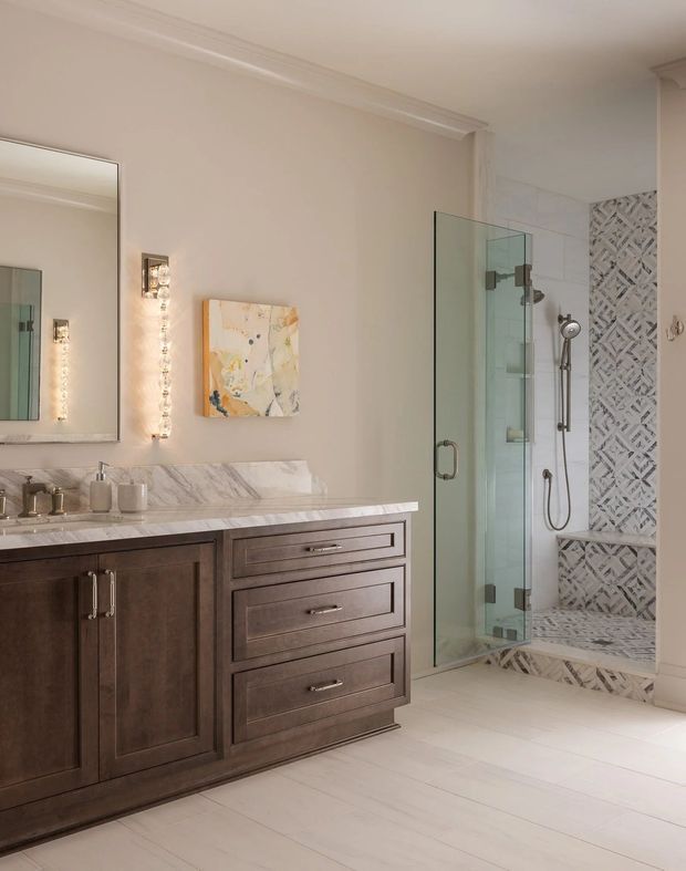 Elegant, bright bathroom featuring a dark stained timber vanity with marble benchtop and backsplash, large framed mirror, wall sconces, and a glass-enclosed shower. The shower includes a handheld fixture, built-in bench, and a patterned marble mosaic feature wall. White marble-look flooring and restrained decorative detailing create a refined traditional aesthetic.