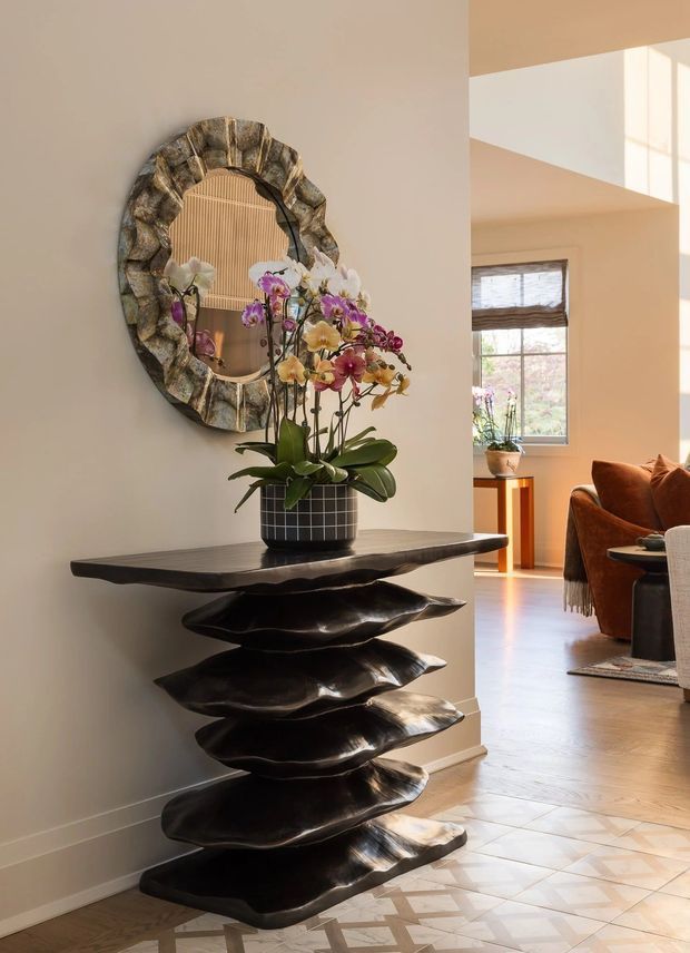 A warmly lit residential entryway featuring a sculptural dark wood console table with layered, organically shaped shelves. An orchid arrangement sits on top beneath a large irregular metallic-framed wall mirror. The entry opens toward a bright living room with timber flooring, a window, and warm brown seating. The foreground floor has a patterned light tile finish.