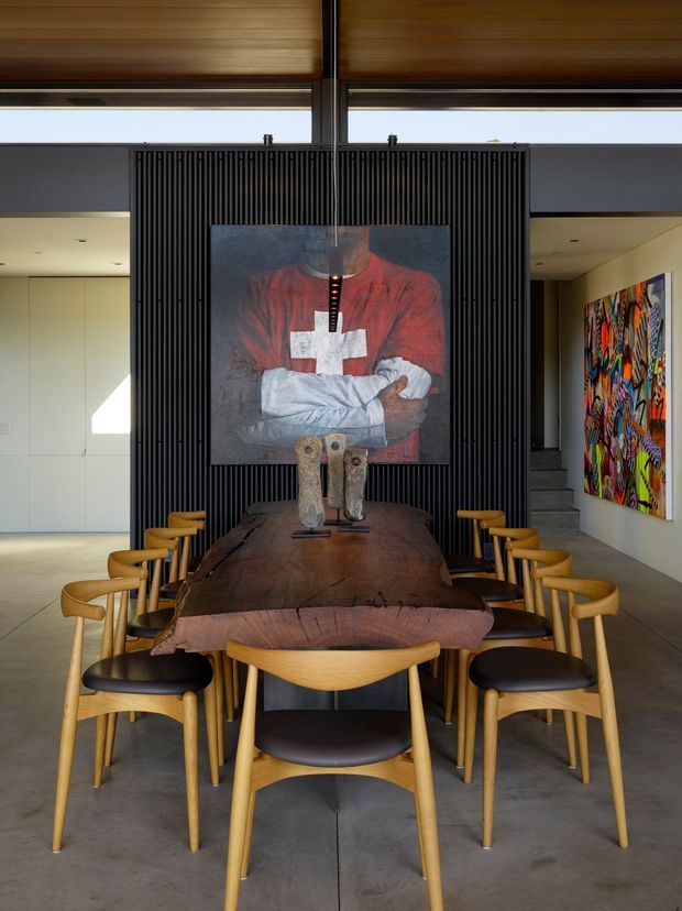 A contemporary dining area with a long, live-edge dark timber dining table surrounded by light timber and black leather chairs. A black vertical-slatted feature screen forms the backdrop, displaying a large artwork, while additional colourful artwork hangs on the right wall. The space has polished concrete flooring, light built-in cabinetry, expansive clerestory glazing, and a warm timber ceiling.