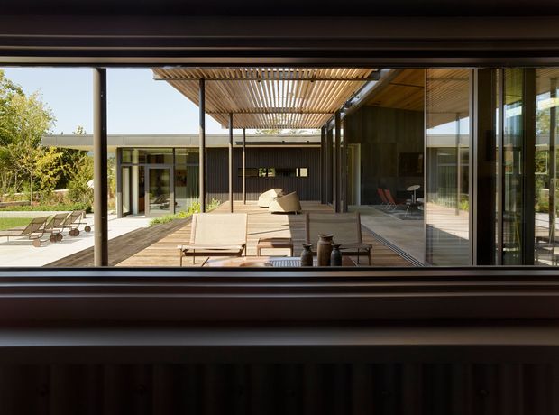 View from inside a dark-framed building through a large opening toward a contemporary courtyard terrace. The scene features timber decking, outdoor lounge furniture, a slatted timber pergola, extensive floor-to-ceiling glazing, dark vertical cladding, and low horizontal roof forms surrounding a landscaped courtyard and poolside area.