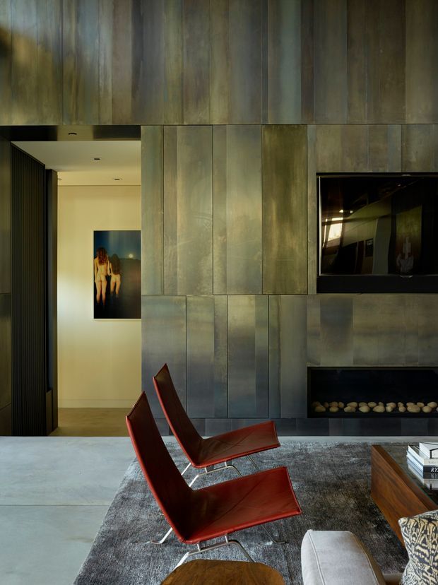 A sophisticated living room with a dramatic full-height metallic wall treatment surrounding a recessed television and linear gas fireplace. Two red leather sling lounge chairs sit on a large grey rug beside a pale sofa and timber coffee table. The space opens to a hallway with artwork, and is lit warmly with a refined contemporary atmosphere.