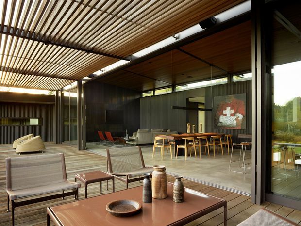 A sophisticated indoor-outdoor living and dining area opening onto a timber deck. The space features extensive floor-to-ceiling glazing, dark ribbed wall cladding, warm timber ceilings and a slatted timber pergola, with a lounge, dining table, and outdoor seating arranged around a central entertaining area.