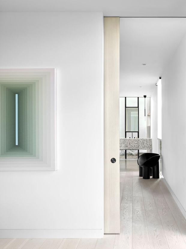 Bright minimalist hallway or entry passage with white walls, pale timber flooring, and a light timber pivot door partially open toward a refined living area. A large abstract framed artwork in soft green and white tones dominates the left wall. Through the opening, a black sculptural chair, terrazzo-patterned partition or counter, black-framed glazing, and a pale upholstered sofa are visible. The space has clean linear detailing, concealed or recessed lighting, and a calm monochromatic palette.