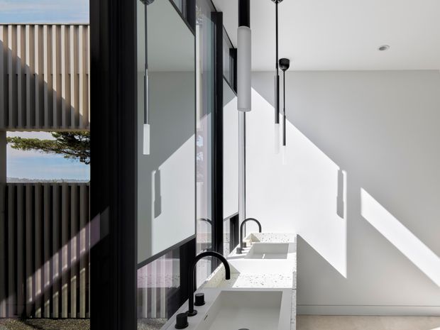 Bright minimalist bathroom with a long white double vanity beneath suspended cylindrical pendant lights. Black-framed full-height glazing and a dark vertical slatted screen line the left side, while strong sunlight creates geometric shadows across the white walls. Speckled white stone benchtops and integrated basins contribute to the clean contemporary fit-out.