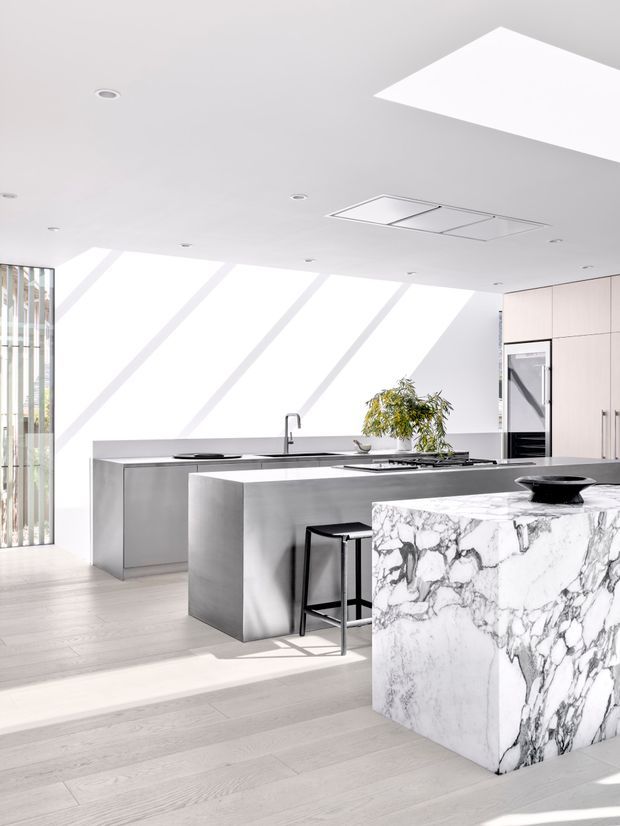 Bright contemporary kitchen with a large marble-clad island in the foreground, stainless-steel cabinetry and secondary island, pale timber flooring, light-toned built-in cabinetry, integrated appliances, and extensive daylight entering through tall glazing. The space has a clean minimalist aesthetic with strong diagonal sun shadows across the white walls.