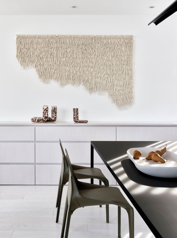 A bright, minimalist dining area with a long dark dining table and sculptural olive-grey chairs in the foreground. Behind it is a full-width light wood built-in cabinet with horizontal drawer fronts, topped by abstract decorative objects. The white wall features a large cream woven textile artwork with an irregular fringed edge, while recessed ceiling lights and strong natural light create a clean, refined atmosphere.