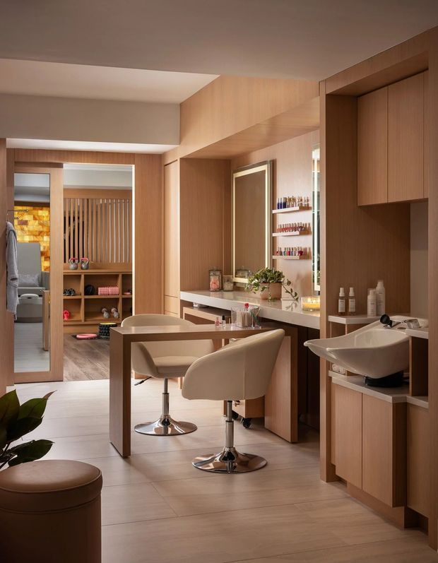 A warm, refined salon or beauty studio interior with extensive light timber built-in cabinetry and wall panelling. Two upholstered styling chairs sit at a central timber workstation facing a large illuminated mirror and countertop. A white ceramic wash basin is integrated into cabinetry on the right, while open shelving, product displays, a waiting stool, and a partially visible treatment area appear in the background. The space has tiled flooring, soft ambient lighting, and a calm neutral palette.
