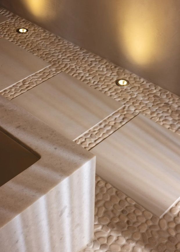Close-up view of a warm, neutral-toned bathroom featuring a rounded stone or plaster vanity edge, pale rectangular wall tiles, pebble-textured feature bands, recessed downlights, and pebble mosaic flooring. The tactile, organic surfaces create a calm spa-like atmosphere.