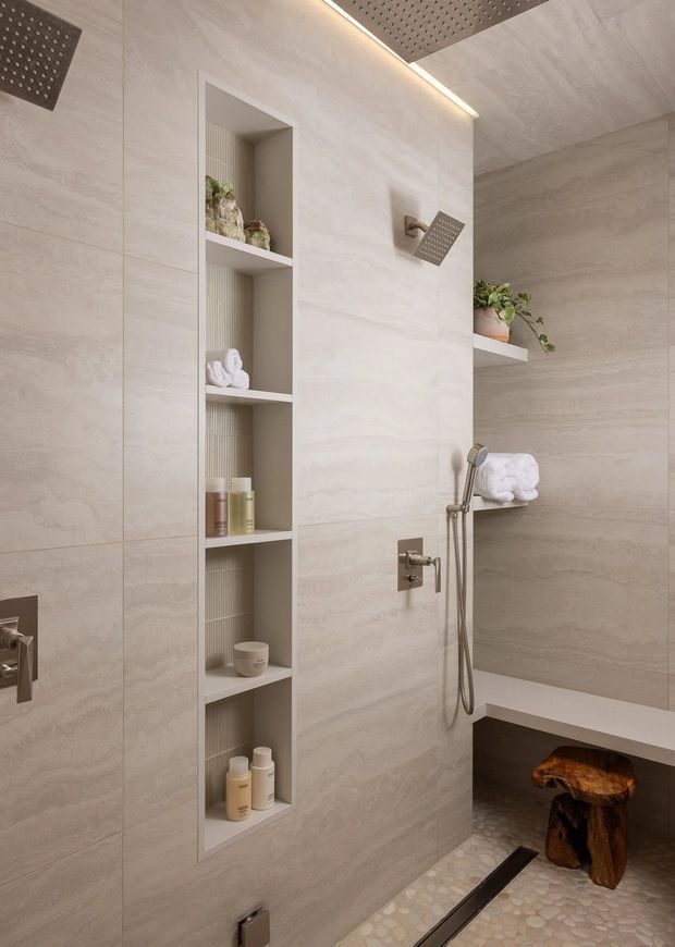 Contemporary bathroom shower area clad in large-format warm beige stone-look tiles, with a built-in open shelving niche, brushed metal shower fittings, recessed ceiling rain shower, floating shelf with plant and towels, and a pebble-style shower floor. A built-in bench and sculptural wooden stool add functional and natural accents.
