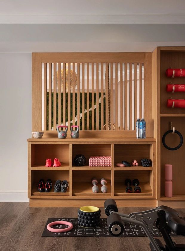 A neatly organised home gym storage wall with warm timber shelving and cabinetry, displaying kettlebells, shoes, weights, resistance equipment and yoga accessories. Vertical timber slats form a screened opening behind the storage, with a view toward greenery beyond. The room has pale walls, white ceiling moulding and dark timber flooring, creating a refined, integrated fitness space.