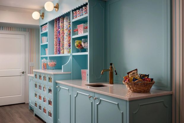 A compact, highly styled pantry or butler's pantry with blue-green painted cabinetry, decorative raised-panel doors, open shelving, and a pale stone benchtop. The shelves display colourful packaged goods and rolls of wrapping paper, while a small sink with a brass tap sits in the counter. The room has white trim, a white panelled door, timber-look flooring, and warm globe wall lights. The design is playful, colourful, and traditional in detailing.