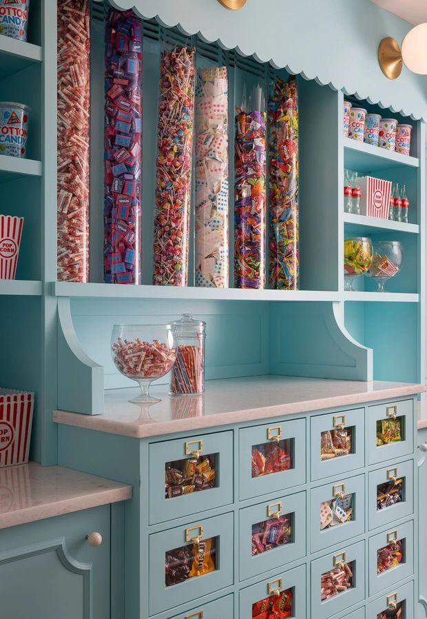A whimsical candy-shop style built-in storage display in a pastel blue room. Tall open shelves hold colourful sweets and lollipops in clear containers, while a pink stone-look countertop sits above a bank of blue drawers with brass label pulls and transparent fronts displaying confectionery. Decorative scalloped trim, curved supports, and wall-mounted globe sconces add a playful, polished finish.