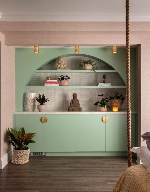 A stylised built-in storage and display wall in a residential interior, featuring a mint-green arched niche with tiled backing, open shelves, decorative objects and plants, and matching lower cabinetry with round brass-toned pulls. The installation is framed by pale textured walls and a substantial horizontal beam, with dark timber flooring and a rope detail in the foreground.