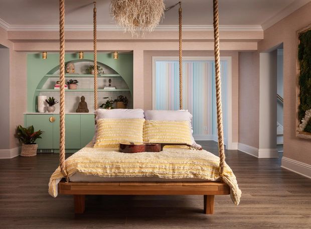 A warm, playful bedroom centered on a suspended wooden swing bed with thick rope supports, layered yellow-and-white bedding, and pillows. The room features dusty pink walls with decorative trim, dark timber flooring, a mint-green built-in arched shelving unit with plants and decor, vertical striped window blinds, and a textured statement pendant light.