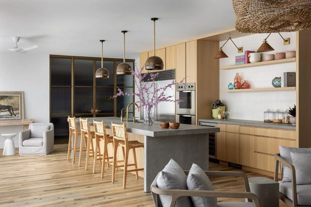 Open-plan kitchen and living area with light timber cabinetry, a large dark stone island with timber bar stools, integrated appliances, open shelving, pendant lighting, and a small seating area. The room has warm timber flooring, neutral upholstery, black-framed glazed doors, and a woven ceiling light.