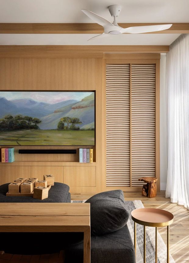 Warm contemporary living room with extensive light timber wall panelling and built-in media cabinetry, a wall-mounted television, dark charcoal sectional seating, and a light timber dining or console table in the foreground. Full-height sheer curtains provide soft daylight beside a tall woven roller blind, while a sculptural white ceiling fan, landscape artwork, books, and a small round metallic side table add detail.