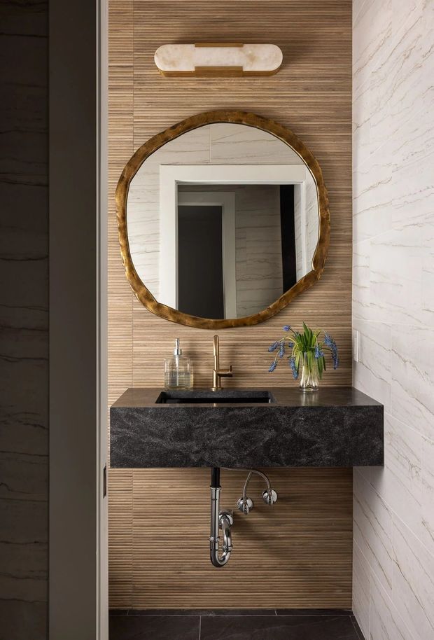 A refined bathroom powder-room vanity with a floating dark stone benchtop, integrated black basin, brushed brass tapware, and exposed chrome plumbing. The vanity wall is clad in warm horizontal timber slats, paired with a large irregular round mirror in a sculptural bronze-gold frame and a decorative wall light. White marble-look wall cladding with grey veining frames the space, with dark floor tiles underfoot.