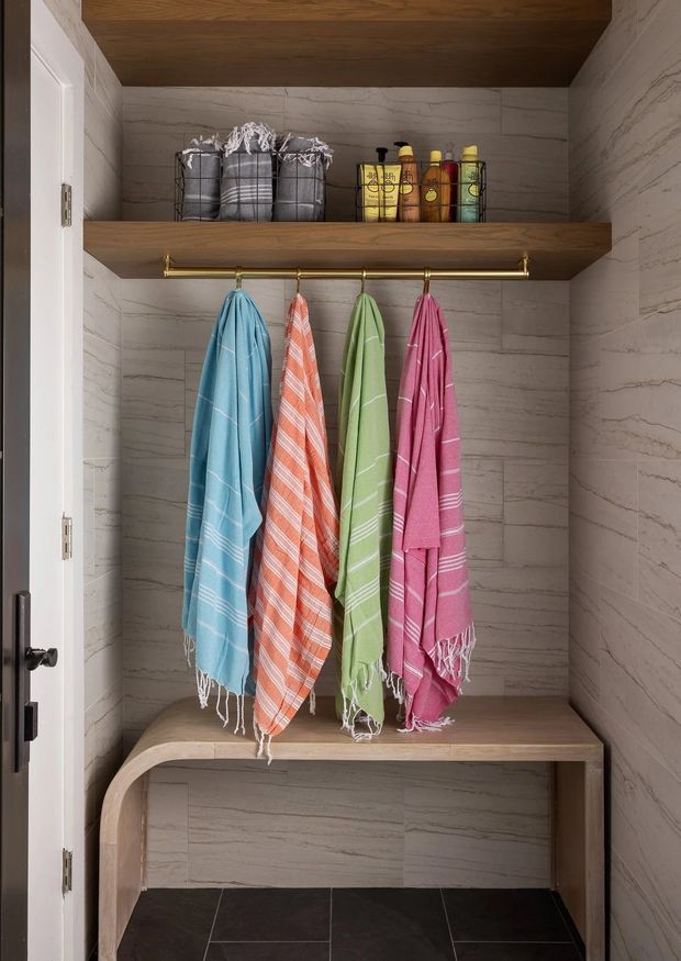 A compact, built-in storage nook or mudroom-style entry area with marble-look tiled walls, dark tiled flooring, warm wood open shelving, a brass hanging rail, colourful towels, baskets and toiletries, and a curved-leg timber bench below.