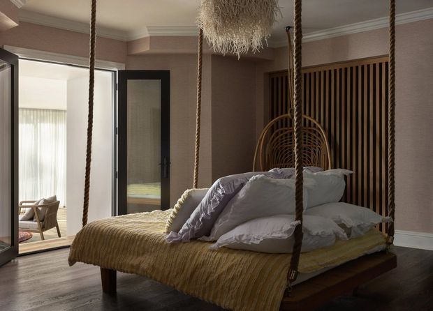 A warm, moody bedroom with a suspended four-poster-style bed hung from thick rope supports. The room features taupe walls with traditional cornice moulding, dark timber flooring, a ribbed timber feature wall, layered white and pale lavender bedding, and a sculptural woven pendant light. Open glazed doors connect to a bright adjoining sitting area.