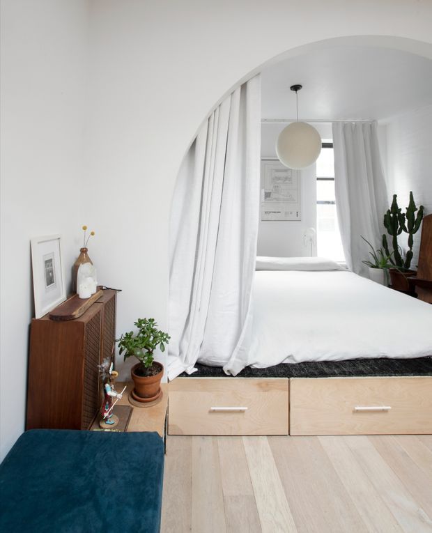 Bright, compact bedroom with a raised platform bed framed by a curved white architectural partition and floor-length curtains. The room features pale timber flooring and integrated pale wood storage drawers beneath the bed, with a dark teal upholstered bench, mid-century wooden sideboard, potted plants, framed artwork, and a spherical pendant light. The overall interior is clean, airy, and softly styled.