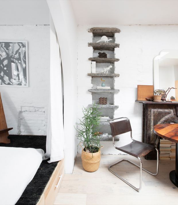 Bright eclectic interior showing a compact bedroom area beside a small workspace. The room features white walls and light timber flooring, a white bed with a black rug, a freestanding dark leather-and-chrome cantilever chair, a timber desk, an ornate dark fireplace mantel, and a rough exposed-brick recessed shelving niche displaying small objects. A potted plant and framed artwork add character.