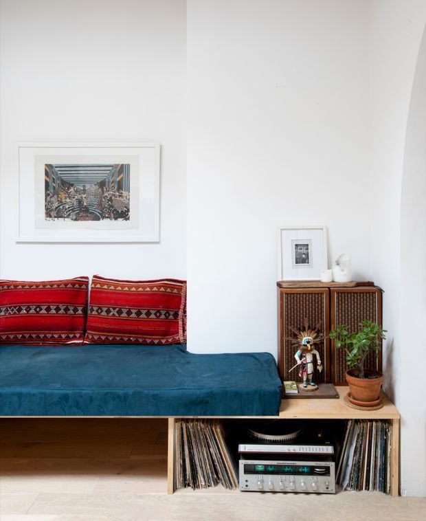 Bright, compact living room corner featuring a deep blue upholstered built-in daybed or sofa with vivid red patterned cushions. A timber media console and open record storage occupy the lower section, with a vintage stereo and vinyl collection. A woven-front timber cabinet, framed artwork, small sculpture, and potted plant add character to the white-walled space.