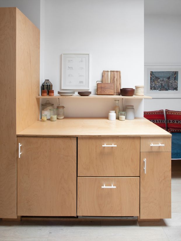 A bright, minimalist kitchen storage and preparation area featuring extensive light timber cabinetry, a broad timber benchtop, open shelving with crockery and jars, and simple white hardware. Framed artwork and a glimpse of a colourful upholstered seating area appear in the background.