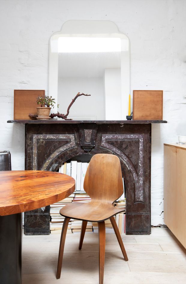 A compact, eclectic dining area featuring a richly veined dark marble fireplace surround with ornate curved detailing, topped by a large rounded rectangular mirror. A warm timber dining table and sculptural wooden chair sit in front, while books fill the fireplace opening and simple timber cabinetry is visible at right. The white textured masonry walls and pale timber floor create a bright backdrop for the dark fireplace and warm wood furnishings.