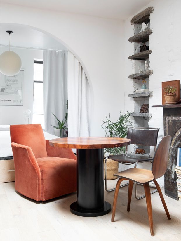 Bright, eclectic living and dining area with a round timber dining table on a black pedestal base, rust-orange upholstered armchair, mid-century style wooden chair, leather chair, indoor plants, and a white-painted brick fireplace wall. A large arched opening leads toward a curtained adjoining area, while rustic exposed masonry shelving provides a strong architectural feature.