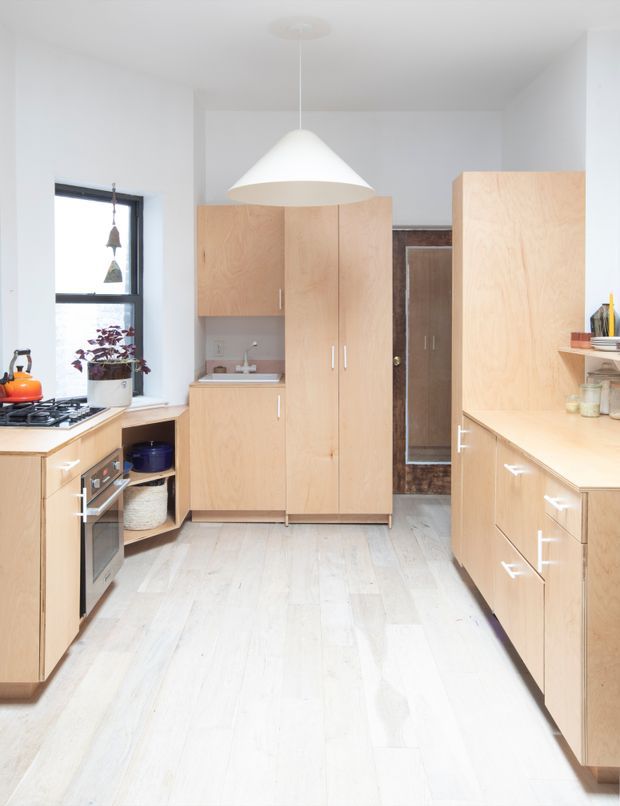 Bright, narrow galley-style kitchen with extensive light timber cabinetry, pale timber flooring, white walls, integrated oven and cooktop, a small sink, and a large white cone pendant light. A window brings in natural light, while open shelving and pantry-style storage are visible along the right side.