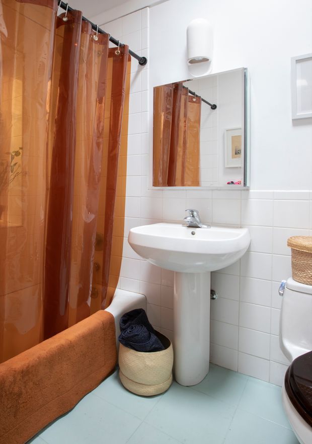 A compact, bright bathroom with white tiled walls, a pale blue tiled floor, a white pedestal sink, bathtub with a translucent amber-brown shower curtain, wall mirror, and toilet. Warm orange and brown textiles and a woven basket add colour and texture.