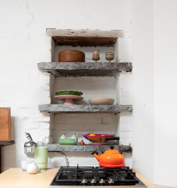 A compact, rustic kitchen vignette centered on an exposed white-painted brick recessed shelving niche. Rough timber and stone shelves display ceramic vessels and serving pieces, while a black gas cooktop and bright orange kettle sit on a pale benchtop in the foreground. The scene combines aged, textured materials with colourful functional kitchenware.