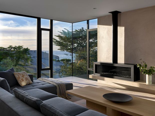 A refined contemporary living room with a large dark sectional sofa, timber coffee table, and a sleek black fireplace with a vertical flue set into a textured feature wall. Floor-to-ceiling corner glazing provides expansive views of trees, neighbouring rooftops, and the ocean beyond. The room has a calm, warm-neutral palette with strong natural light and a connection to the coastal landscape.