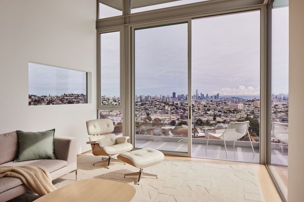 A bright, minimalist high-rise living room with floor-to-ceiling glazing and expansive city views. The room includes a pale upholstered sofa, a modern lounge chair with ottoman, a low light-wood coffee table, and a small outdoor terrace visible beyond the glazing. Neutral finishes and abundant daylight create an airy, refined atmosphere.