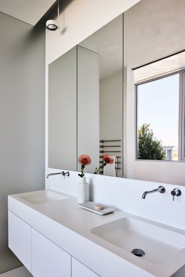 Bright, minimalist contemporary bathroom featuring a long white double vanity with integrated basins, wall-mounted chrome tapware, large frameless mirrored panels, pale tiled walls, and a large window. A towel warmer and subtle decorative styling are visible in the mirror reflection.
