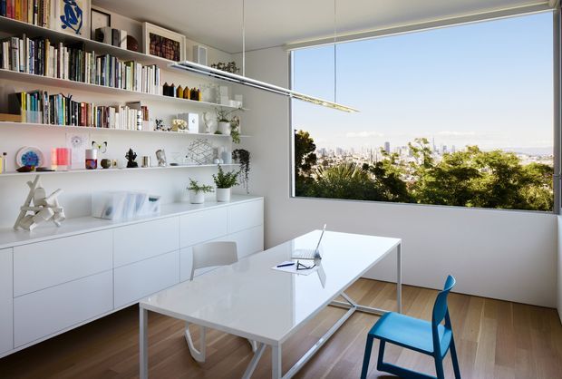 Bright, minimalist home office with extensive white built-in shelving and low cabinetry, a white desk, contemporary chairs, and a large picture window overlooking a leafy urban landscape. The room has light timber flooring, white walls, clean-lined furnishings, and a slim suspended linear light.