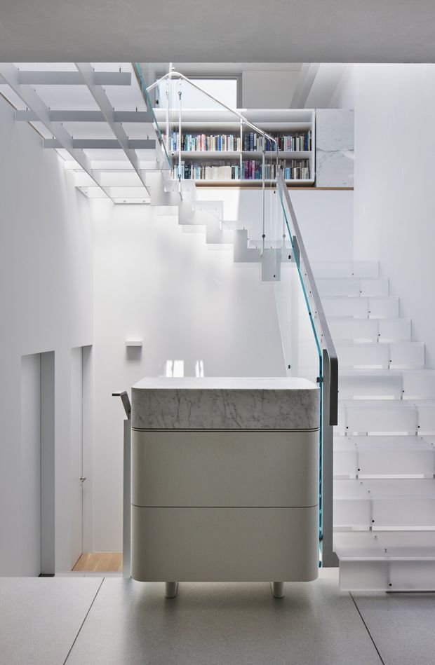 Bright minimalist interior featuring a sculptural white staircase with clear glass and slender metal balustrades, wrapping around a central light-grey kitchen island or built-in cabinet with a marble-look stone benchtop. A built-in bookshelf occupies the upper landing, while white walls, exposed stair structure, skylight-like glazing, and pale flooring create a highly architectural, airy space.