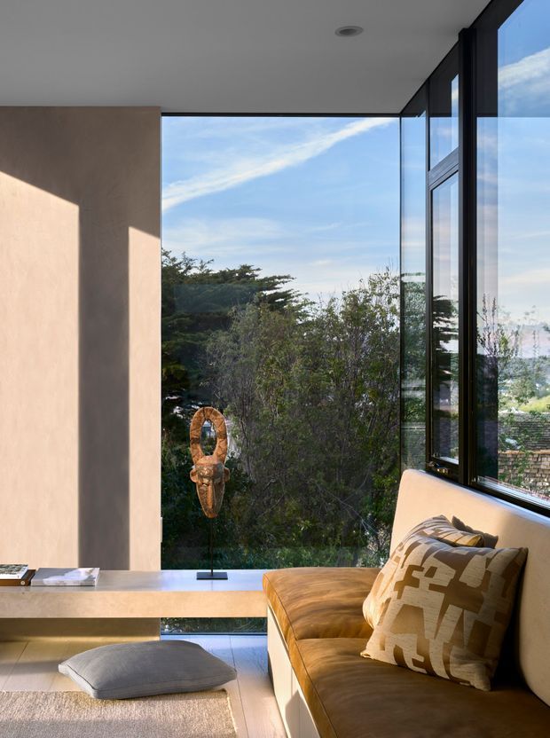 A refined modern living room with a built-in tan leather banquette positioned beside expansive floor-to-ceiling corner glazing. The room has smooth warm beige plaster walls, a pale floor, a slim built-in console, minimalist recessed ceiling lighting, and a sculptural decorative object displayed in front of the windows. Large windows frame leafy vegetation and distant landscape views, while a patterned neutral cushion adds texture to the seating.