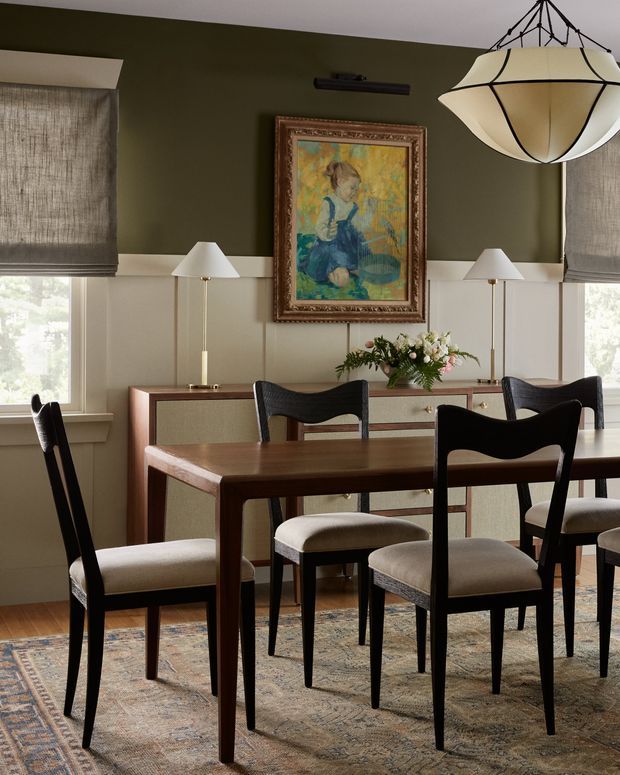 A refined dining room with a dark timber dining table and sculptural black dining chairs upholstered in light beige fabric. Olive-green walls are paired with white wainscoting, framed artwork, matching slim table lamps, and a large sculptural pendant light. A vintage-style patterned rug, timber sideboard, floral arrangement, and linen Roman blinds complete the warm, traditional setting.
